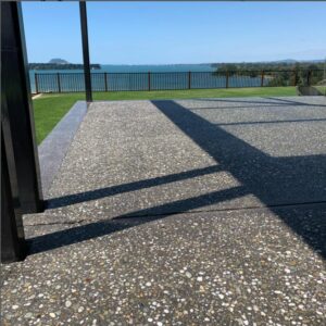 1 an outside pastio photo showing a bush hammer exposed aggregate concrete floor with a large grass lawn and the sea in the background and a striped fence at the edge of the property beyond the grass