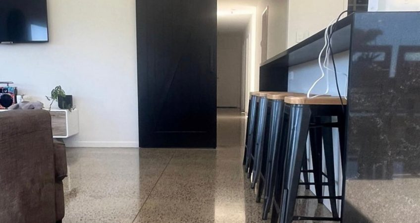 an open plan kitchen diner with the focus on a polished concrete floor, a barn door is in the background painted black and a sofa is to the left, a black kitchen bench to the right with 4 barstools